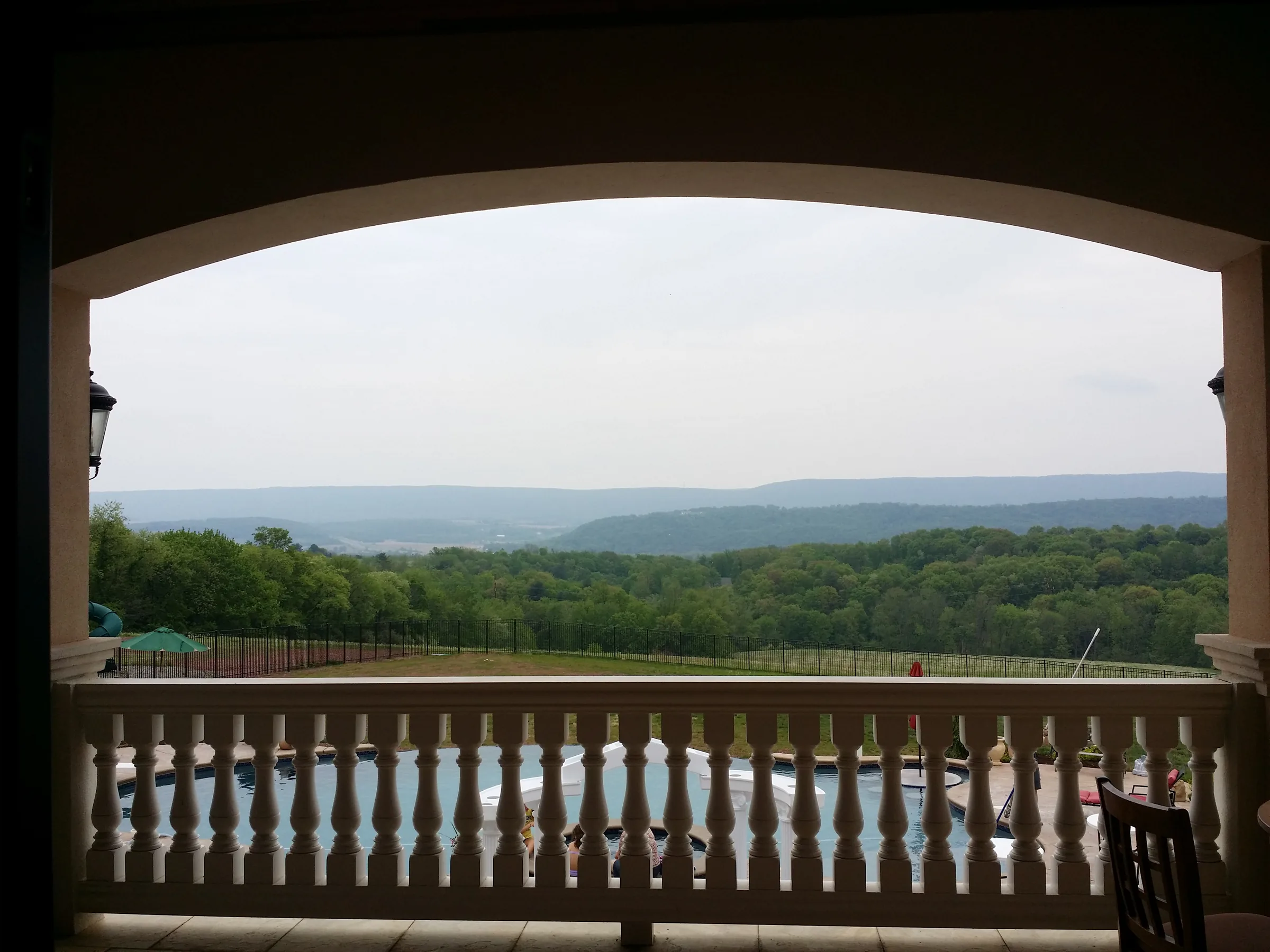 View from a covered balcony through an arched opening, overlooking a pool and wooded mountain valley