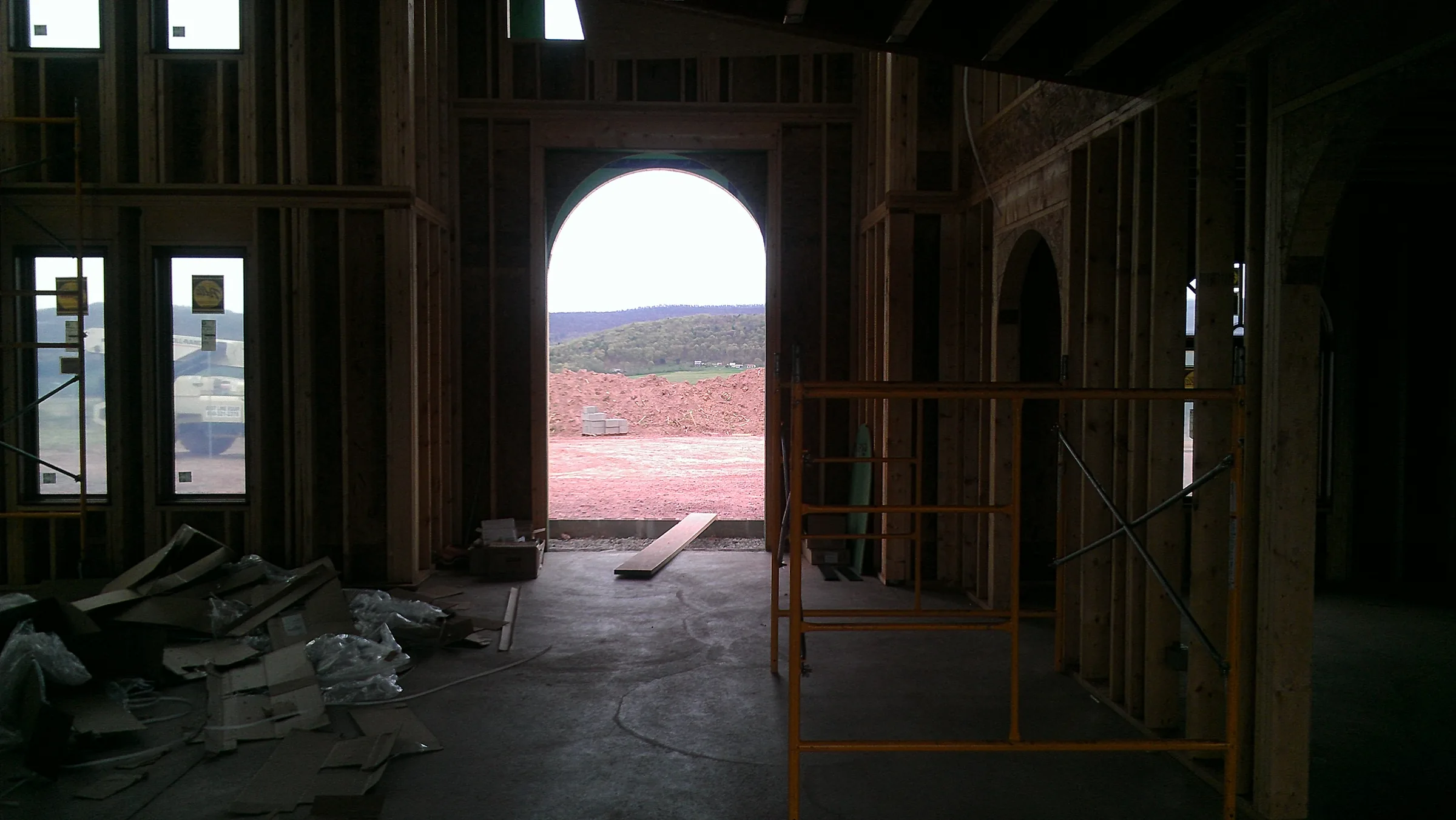 View through a framed arched entry opening during rough construction, with scaffolding and the graded site visible beyond
