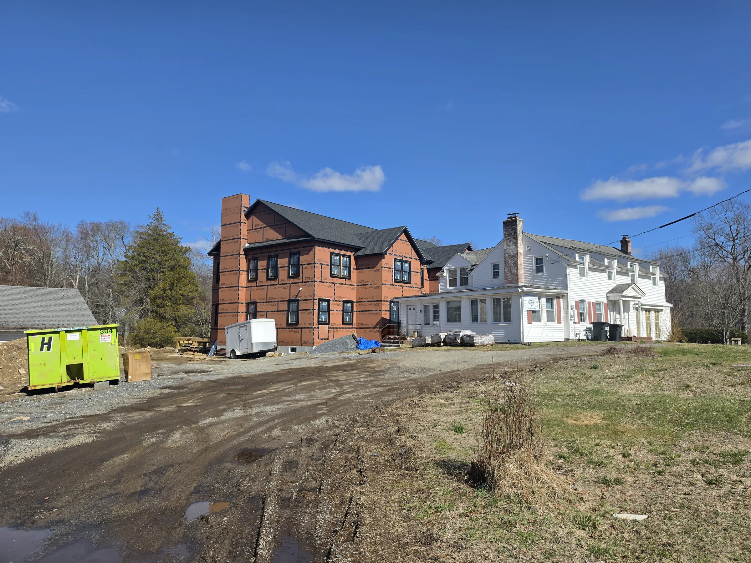 Two-story addition wrapped in brick-patterned weather-resistive barrier, attached to an existing white clapboard house, with a construction dumpster in the dirt yard