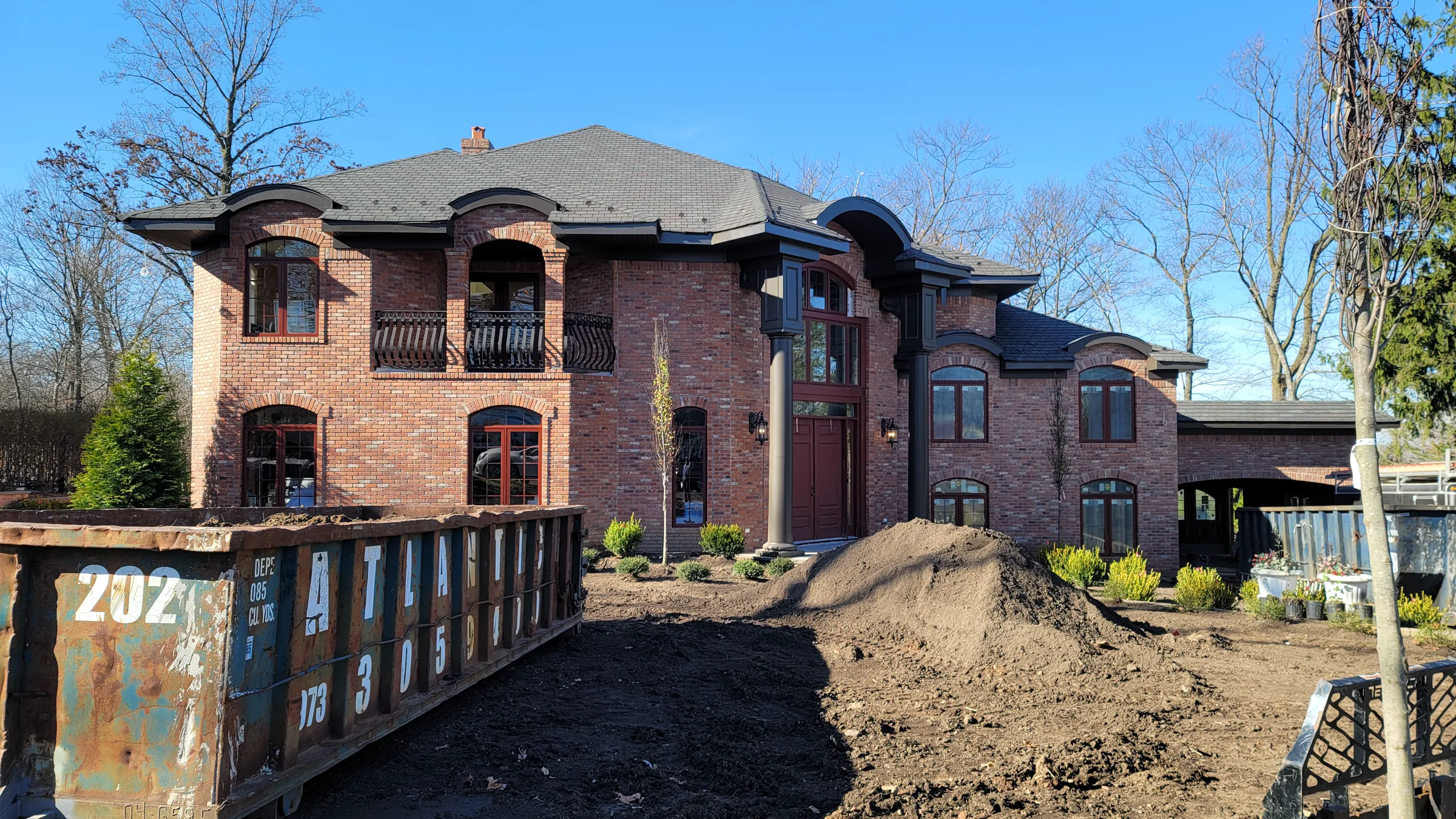Red brick house with a two-story columned entry, arched second-floor balcony bays, and a hipped shingle roof, a roll-off dumpster and mounded soil in the unlandscaped yard