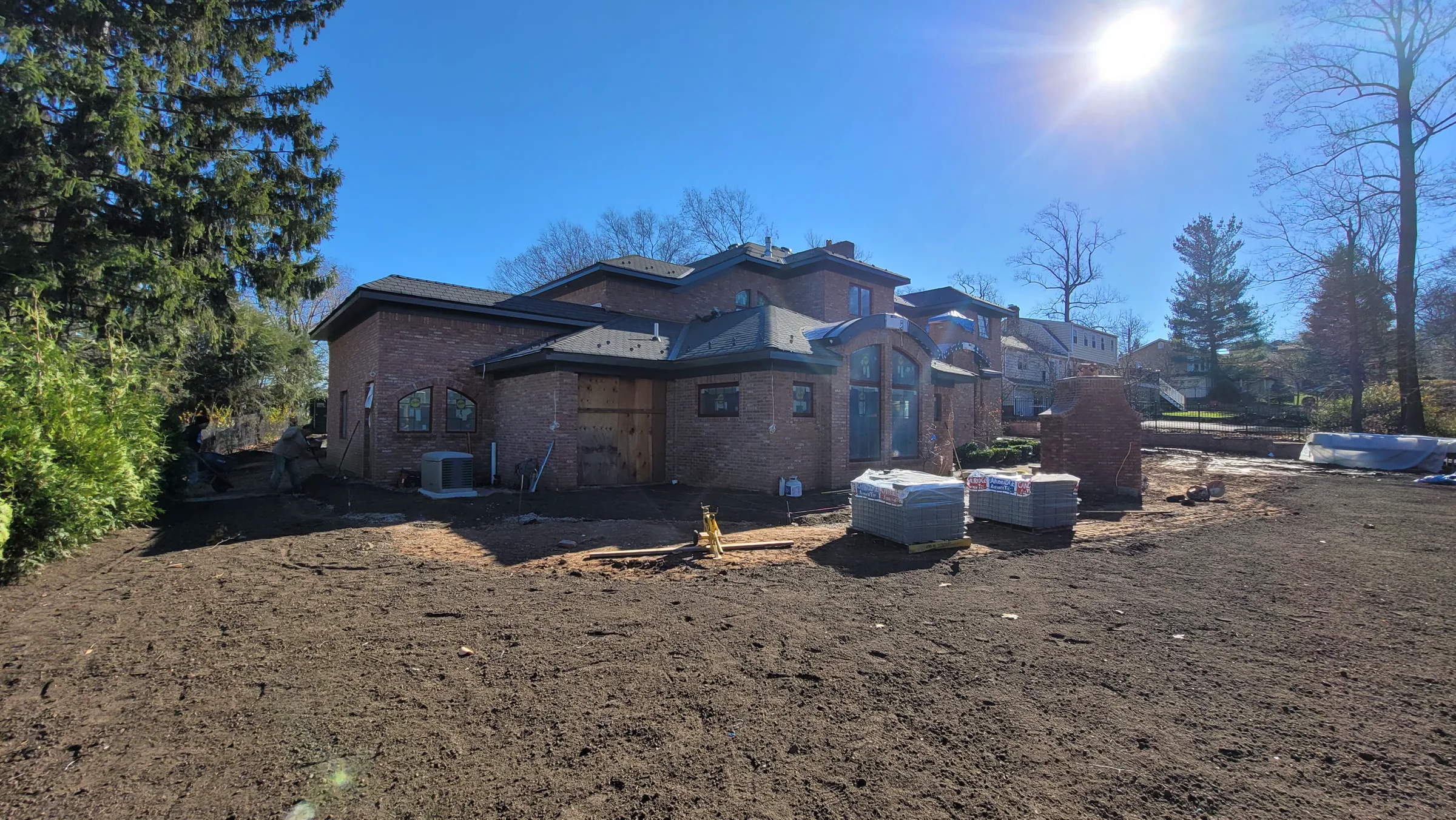 Side and rear elevations of the brick house with a plywood-boarded rough opening, a condenser unit, and banded pallets of material staged on the dirt yard