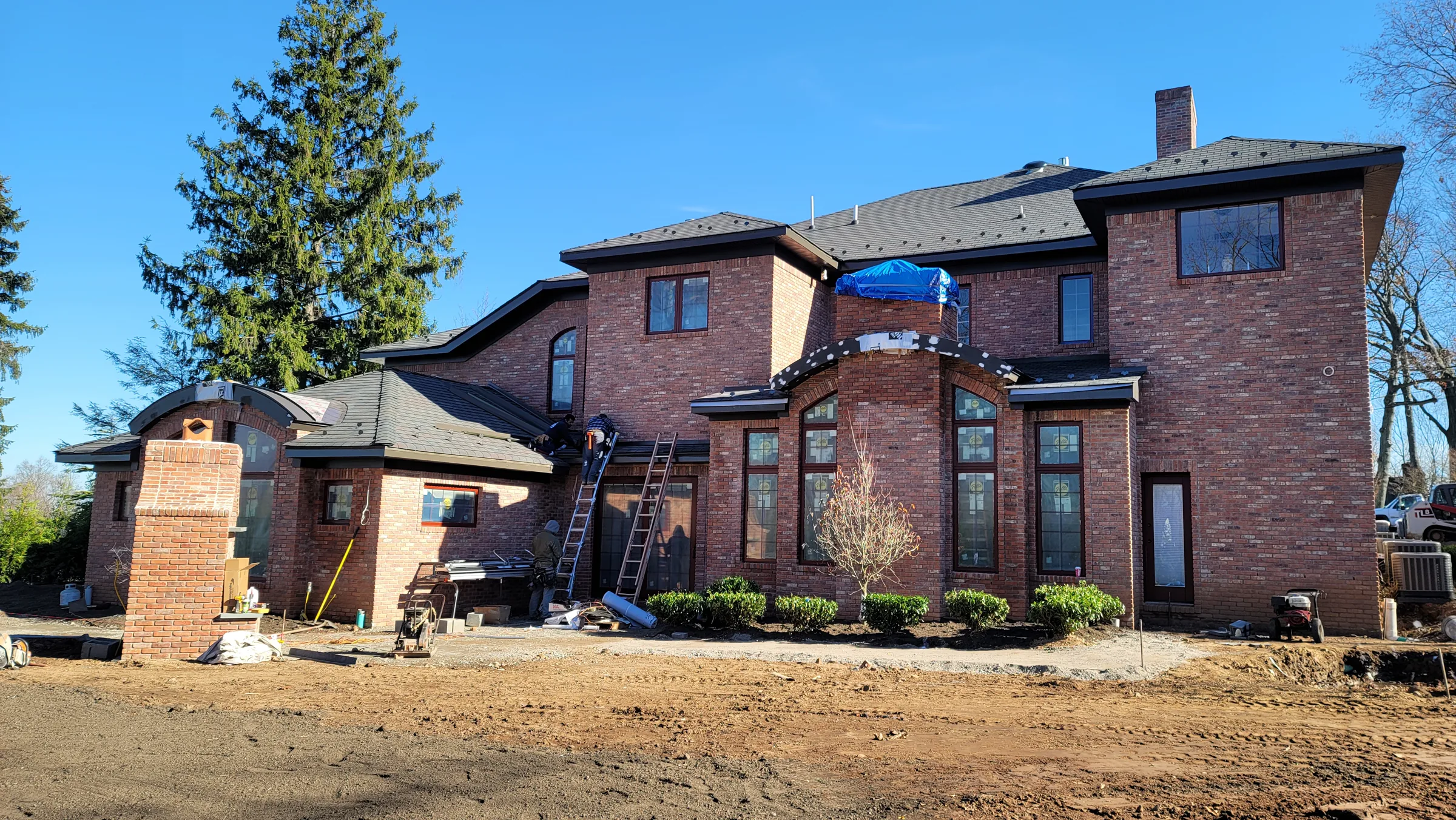 Rear elevation of the brick house with a tall arched window bay, a blue tarp over part of the roof, and an extension ladder set against the wall