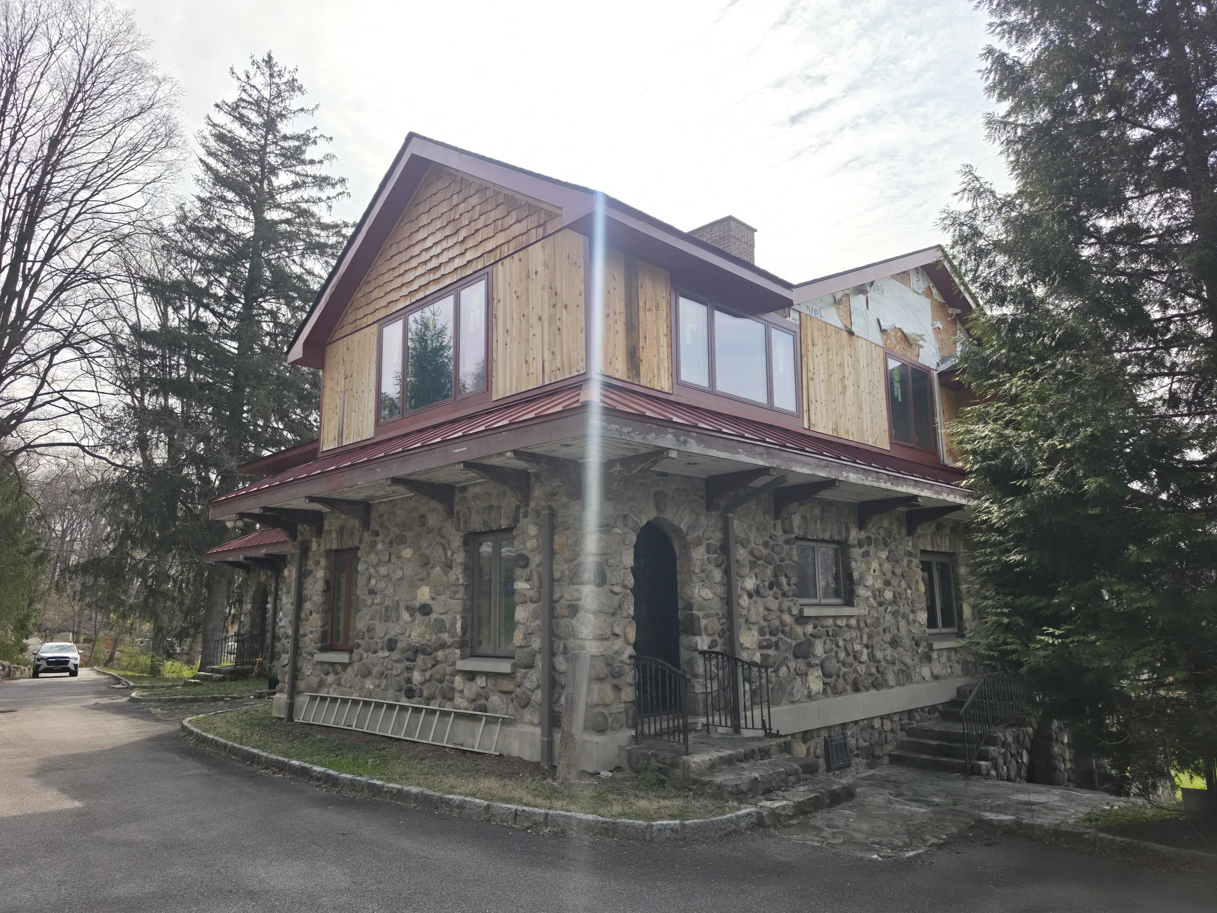 Three-quarter view from the paved driveway of the cedar-clad upper story and red standing-seam porch roof carried on the round river-stone arcade