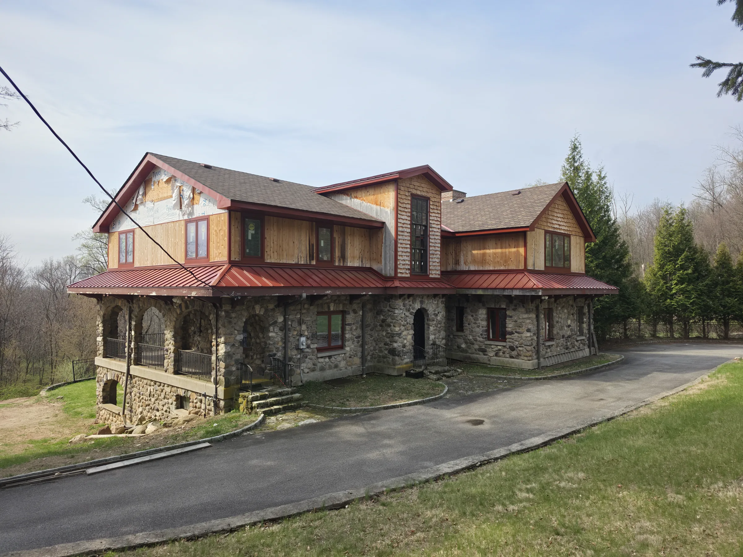 Wide view of the house under construction showing the new cedar cross-gable, red standing-seam roofs, a stone chimney, and the shingled rear wing above the stone ground floor