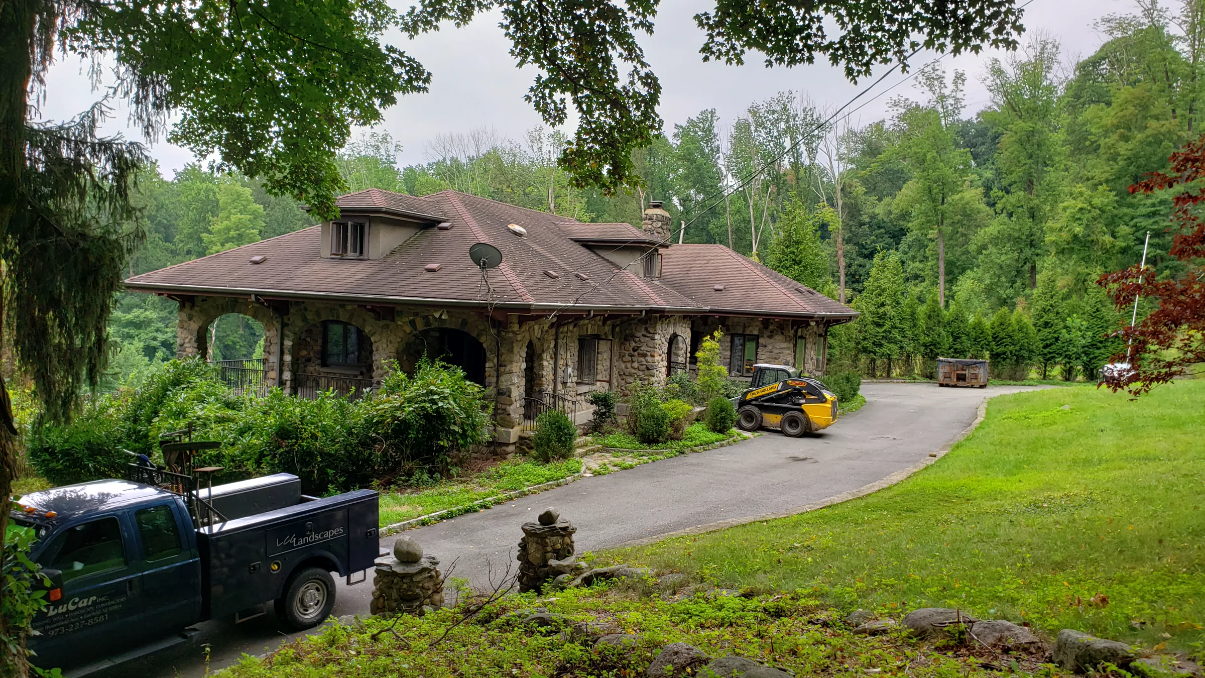 The same river-stone house from nearer the drive, with a landscaping pickup truck and a skid-steer loader parked along the circular driveway