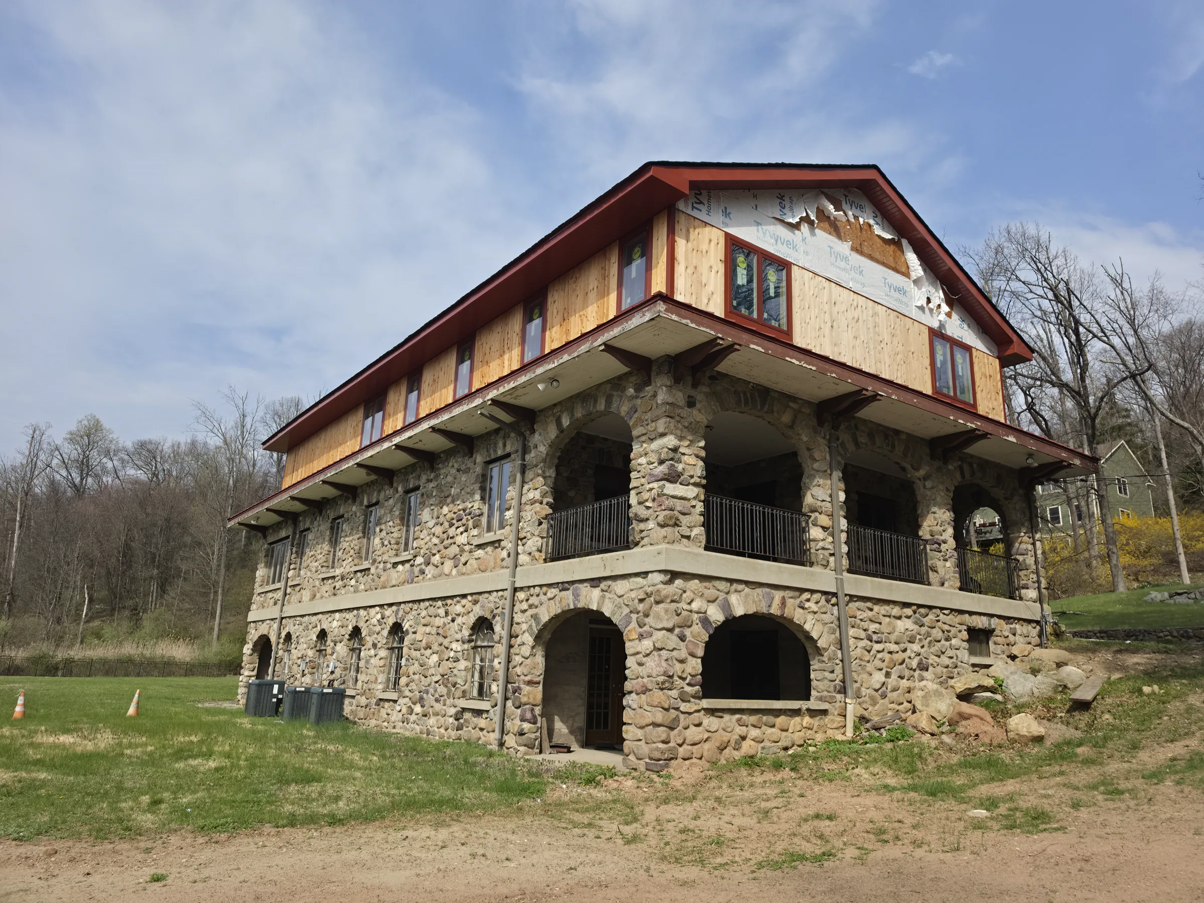 Long river-stone facade with a run of arched openings and a stone-columned balcony, the cedar-clad upper story and red metal roof above