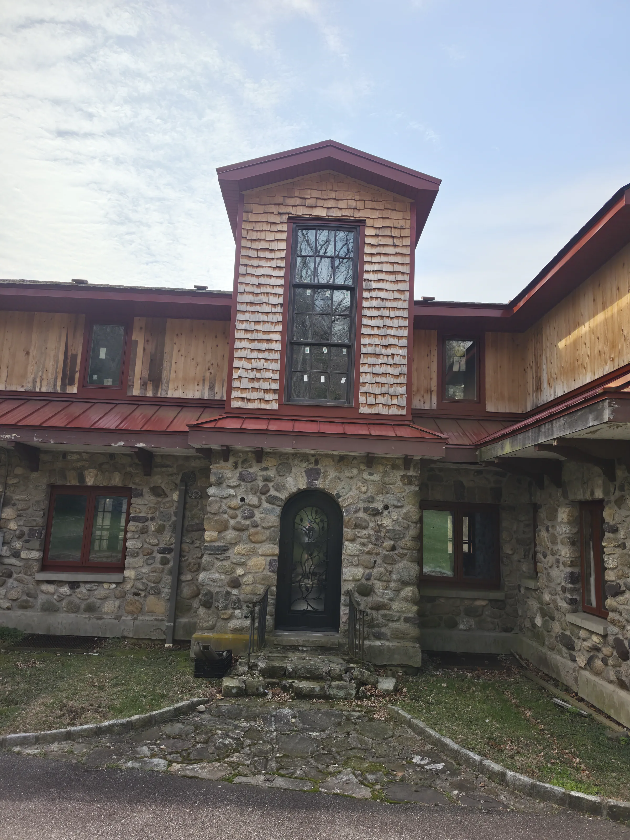 Cedar-shingled entry tower with a tall multi-pane window and red trim rising above a round river-stone base with an arched opening and a dark iron-and-glass door