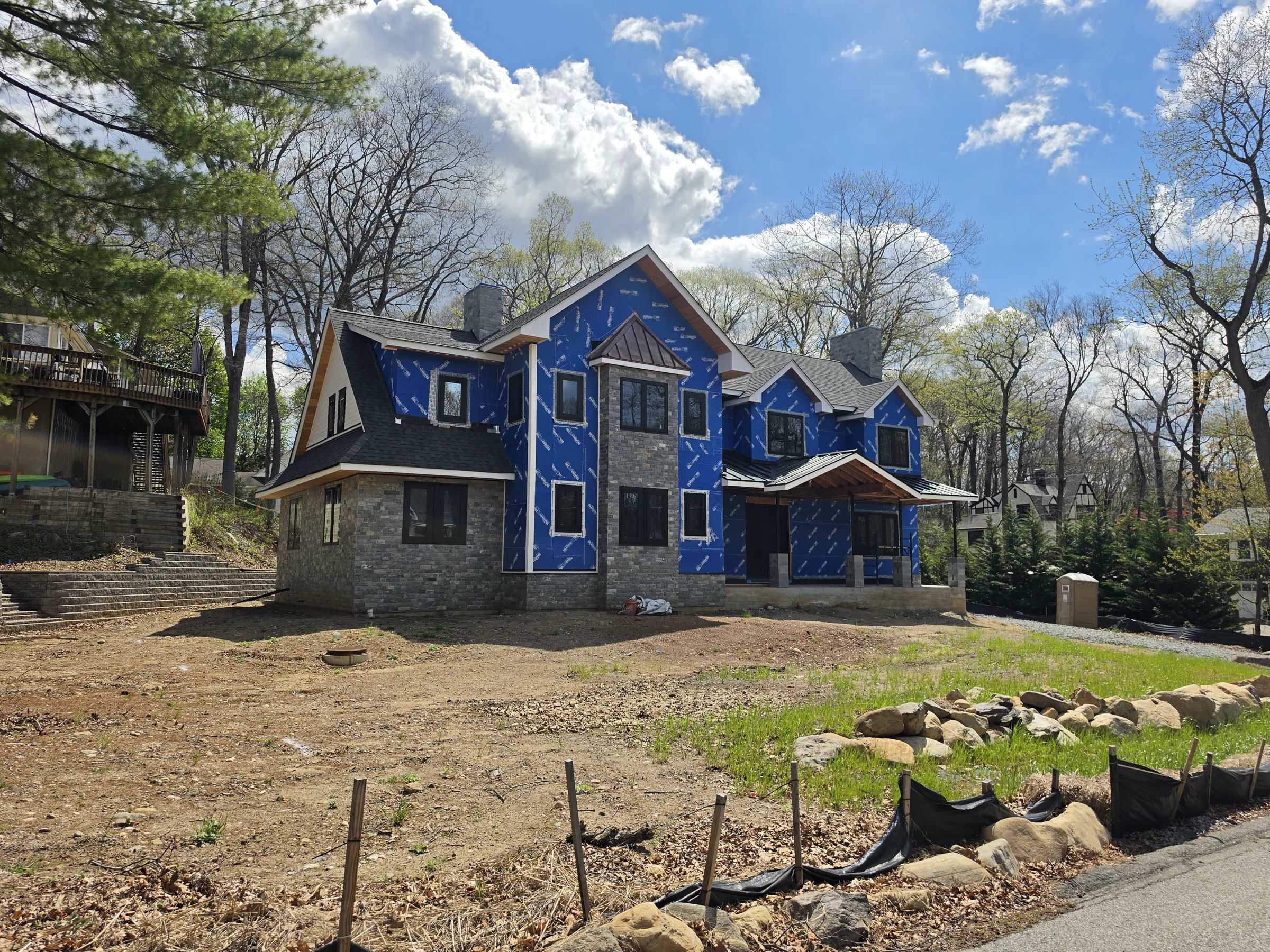 New house under construction, its walls sheathed in blue weather-resistive barrier above a stone-veneer base, with several gables and two stone chimneys