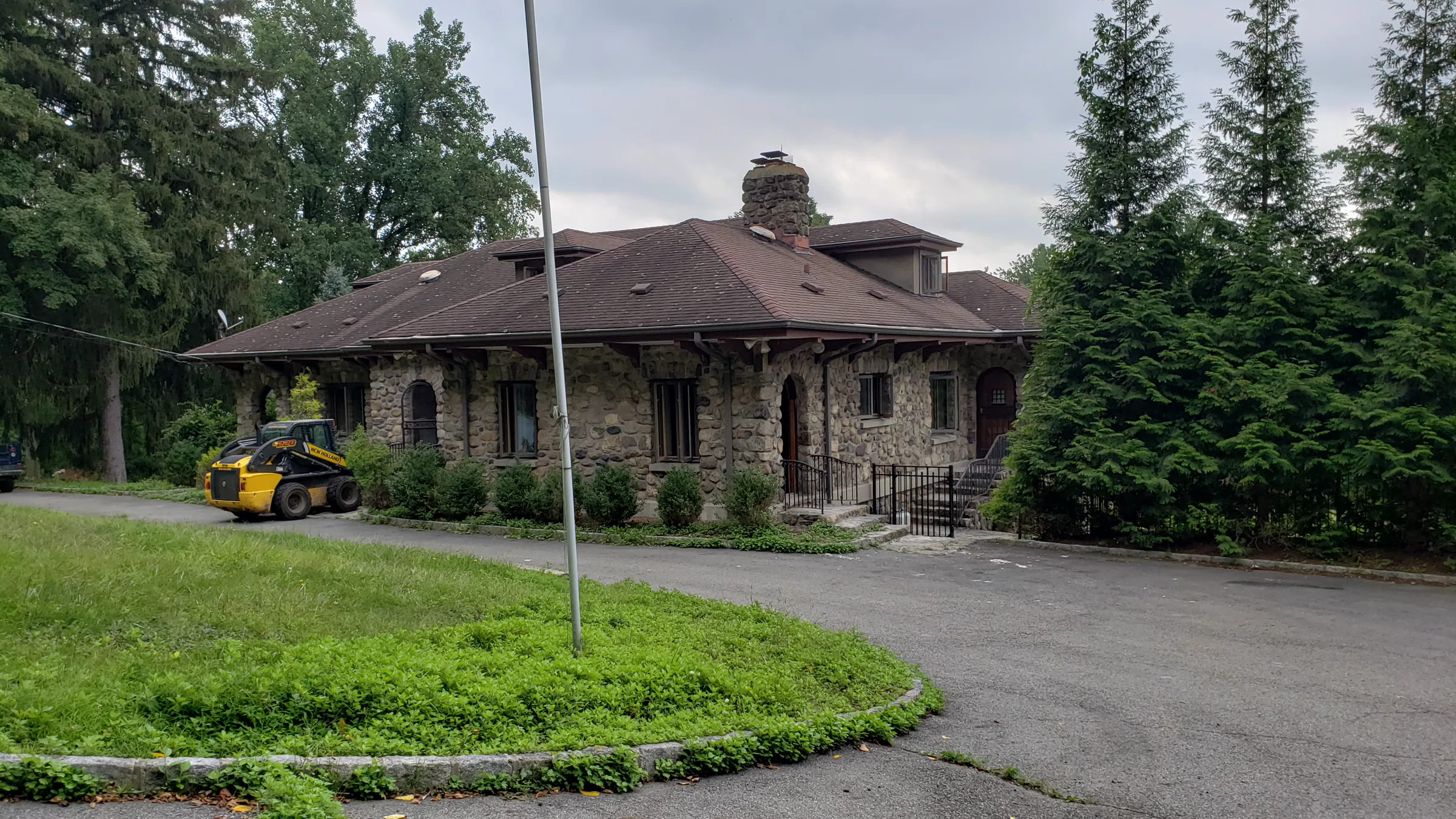 One-and-a-half-story river-stone house with a three-arch stone porch and a hipped shingle roof, seen from the driveway on an overcast day
